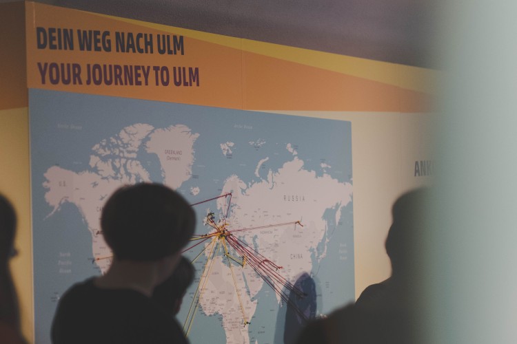 Foto zeigt eine Power-Ppint-Präsentation im Hintergrund, darauz zu sehen eine Landkarte und der Titel "Dein Weg nach Ulm, Your Journey to Ulm"
