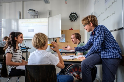 Schüler*innen sitzen um einen Tisch und lachen, reden. Sie sitzen auf Stühlen oder in einem Rollstuhl