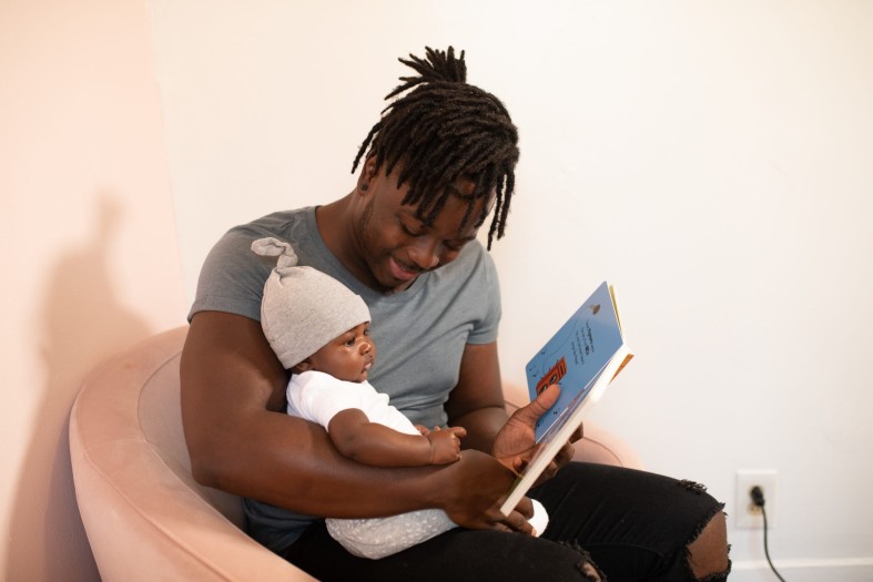 Mann (BiPOC) mit Baby auf dem Arm liest ein Buch