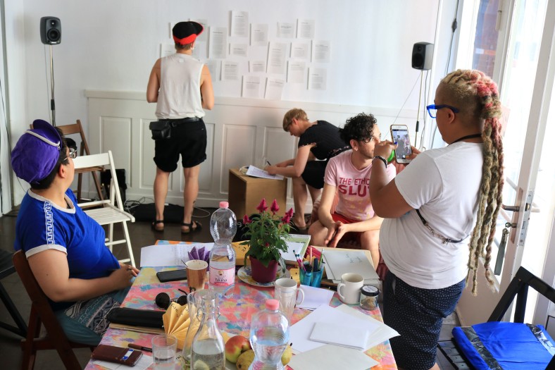 Mehrere Menschen bei einem Workshop, es liegt Papier auf einem Tisch, Zettel mit Informationen hängen an einer Wand, es gibt Getränke