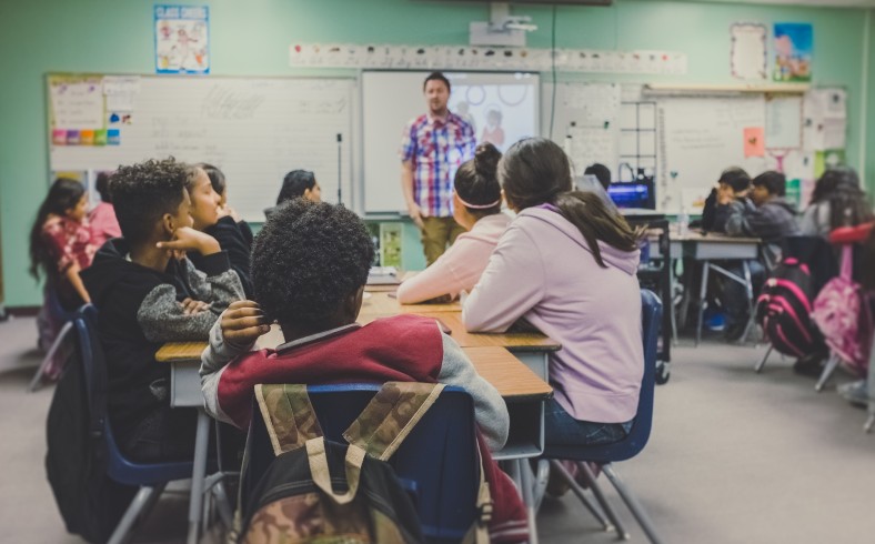 Szene in einem Klassenzimmer, der Lehrer steht vor der Tafel, die Schüler*innen hören ihm zu